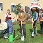 Vier Personen posieren vor einer Brauerei mit Schaufeln, einem Baum, Äpfeln und Gartengeräten auf einer Wiese.
