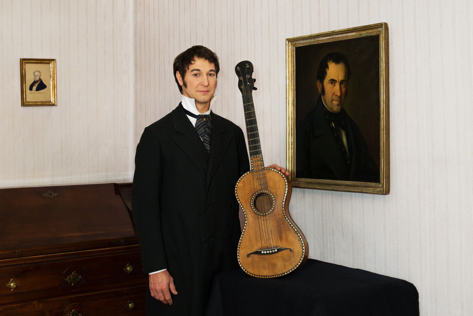 Ein Mann in historischer Kleidung steht neben einer antiken Gitarre und einem gerahmten Porträt an der Wand in einem historischen Raum.