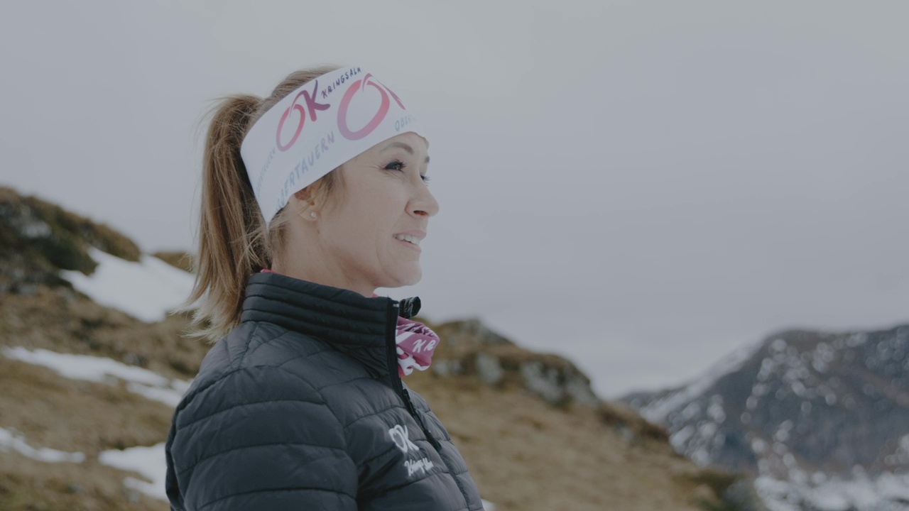 Frau in Wintersportkleidung und Stirnband steht im Freien mit verschneiten Bergen im Hintergrund.