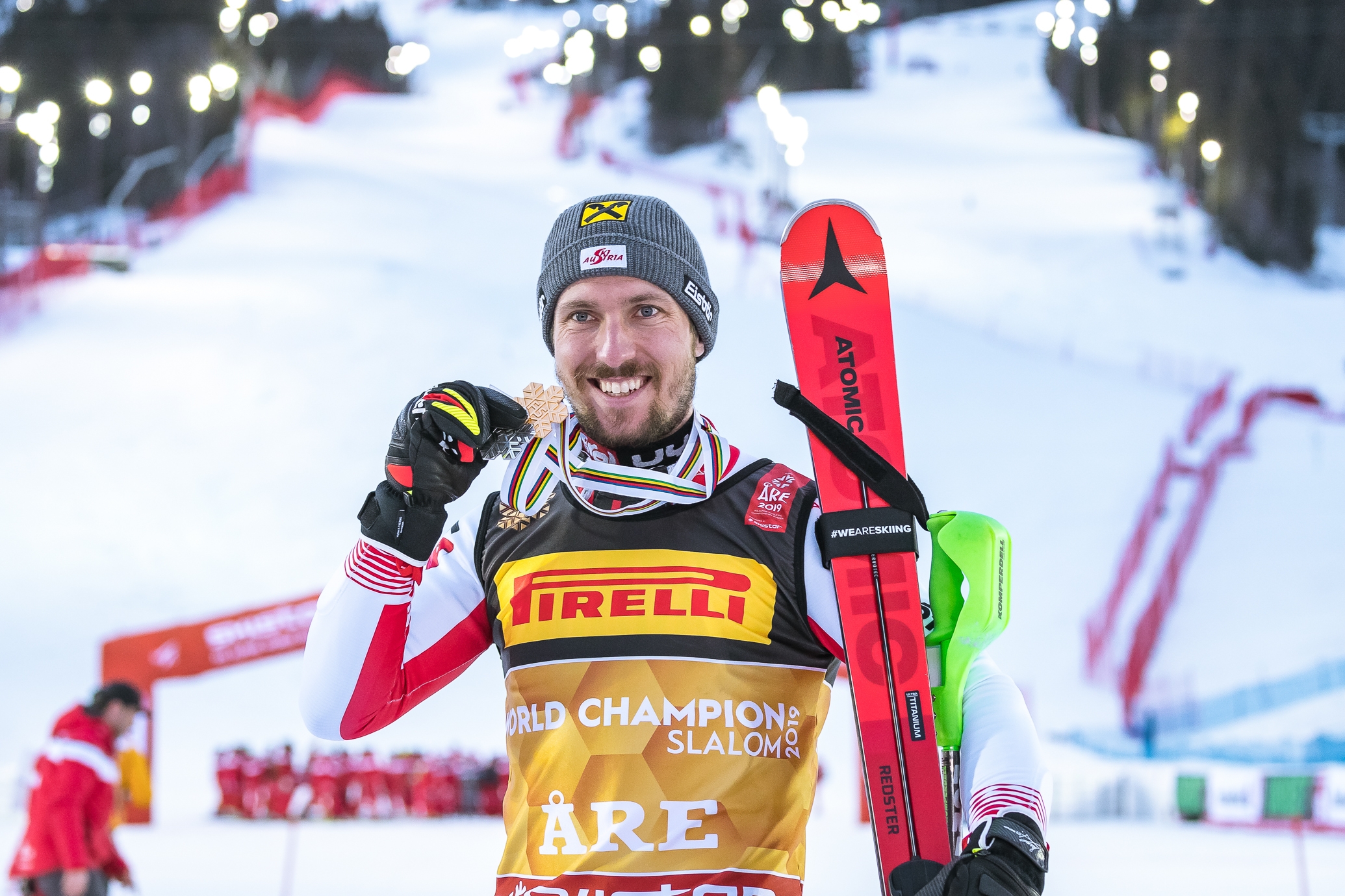 Ein Skifahrer in Rennkleidung lächelt, hält eine Medaille und Skier in der Hand, im Hintergrund eine verschneite Piste und Lichter.