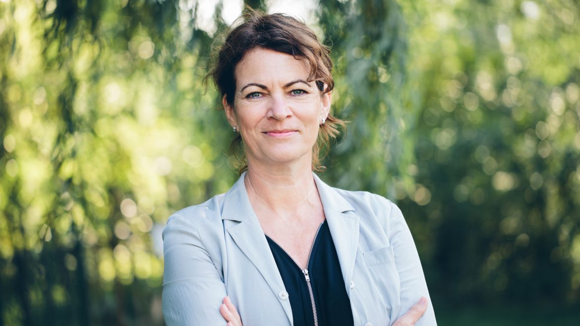 Frau mit kurzen braunen Haaren in einem hellen Blazer steht mit verschränkten Armen im Freien, im Hintergrund Grünpflanzen.