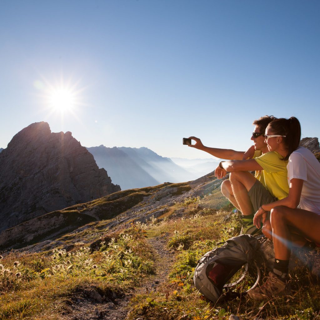 Zwei Wanderer sitzen bei Sonnenaufgang an einem Berghang und machen ein Selfie mit einem felsigen Gipfel im Hintergrund.