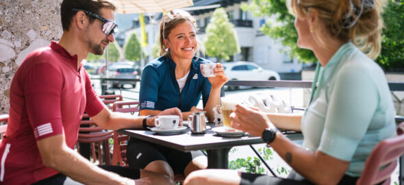 Drei Radfahrer in Trikots unterhalten sich an einem sonnigen Tag in einem Straßencafé und genießen einen Kaffee.