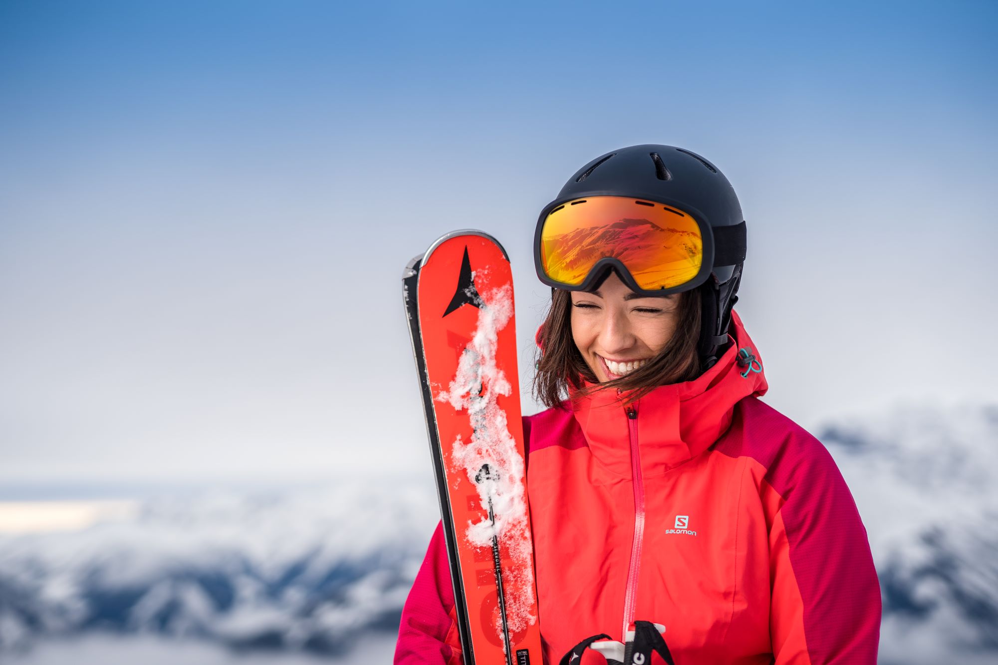 Frau in roter Skijacke und Helm, lächelnd, mit Skiern in der Hand, mit verschneiten Bergen im Hintergrund.