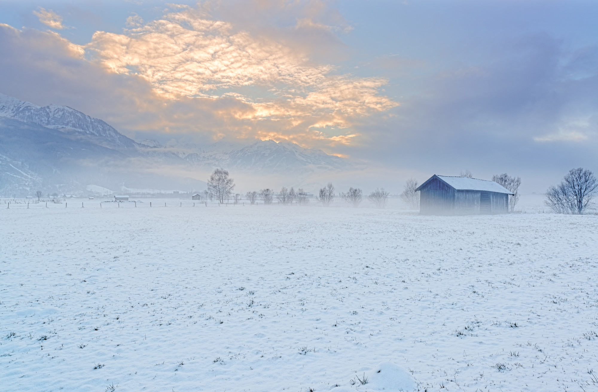 Ein schneebedecktes Feld mit einer kleinen Scheune, Bäumen und Bergen unter einem wolkenverhangenen Himmel bei Sonnenaufgang.