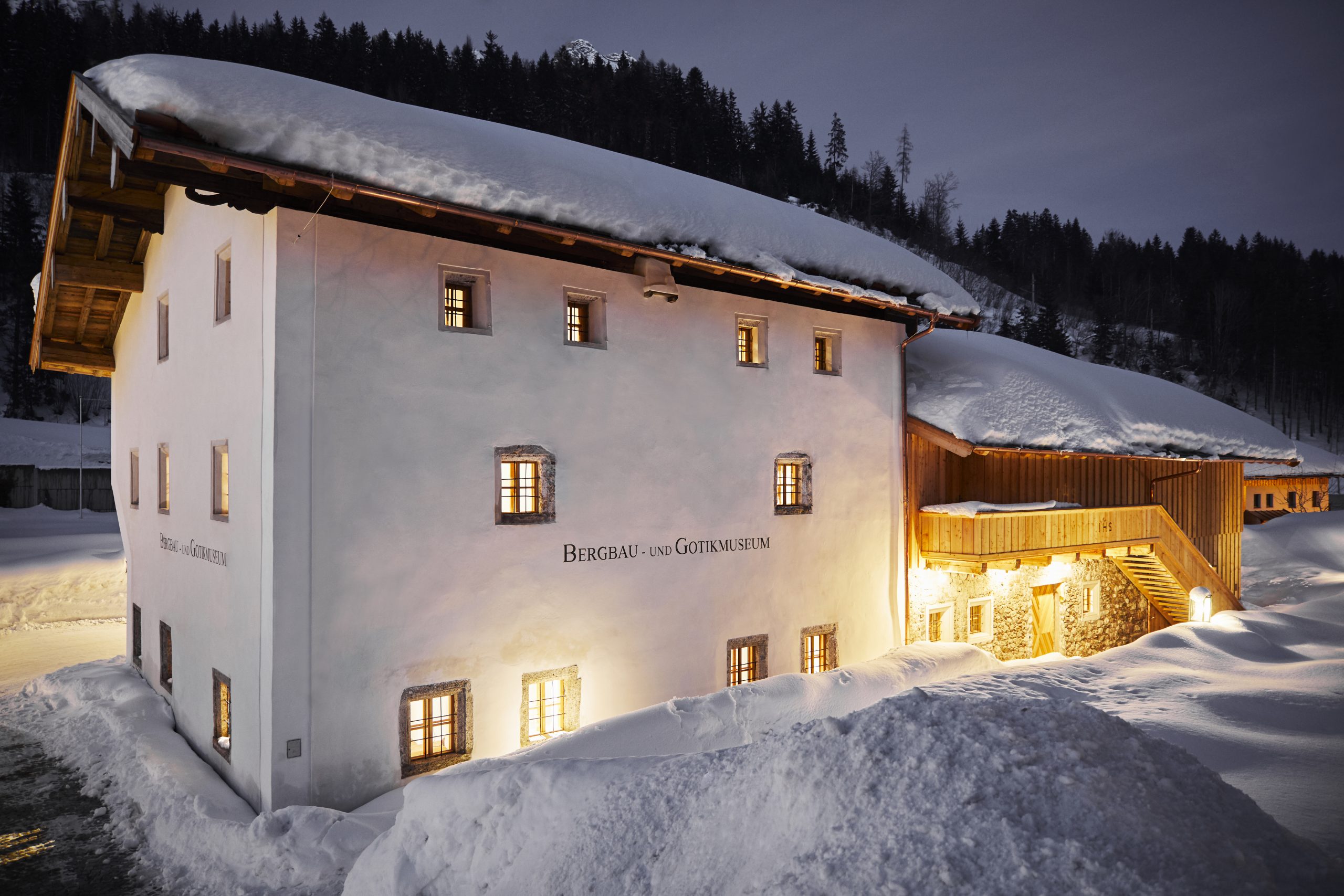Ein schneebedecktes Museumsgebäude leuchtet nachts warm in einer verschneiten, bewaldeten Berglandschaft.