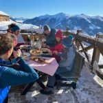 Eine Familie genießt eine Mahlzeit im Freien auf einer verschneiten Berghütte mit malerischem Bergblick.