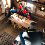 Fünf Personen sitzen um einen Holztisch in einer gemütlichen Hütte, durch deren Fenster das Sonnenlicht fällt.