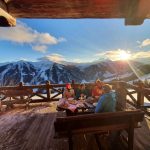 Menschen essen im Freien auf einer verschneiten Bergterrasse bei Sonnenuntergang mit malerischem Alpenblick im Hintergrund.