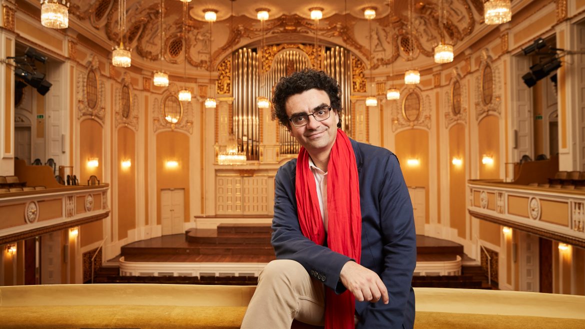 Ein Mann mit blauem Blazer und rotem Schal sitzt in einem prunkvollen Konzertsaal mit Kronleuchtern und einer großen Pfeifenorgel.