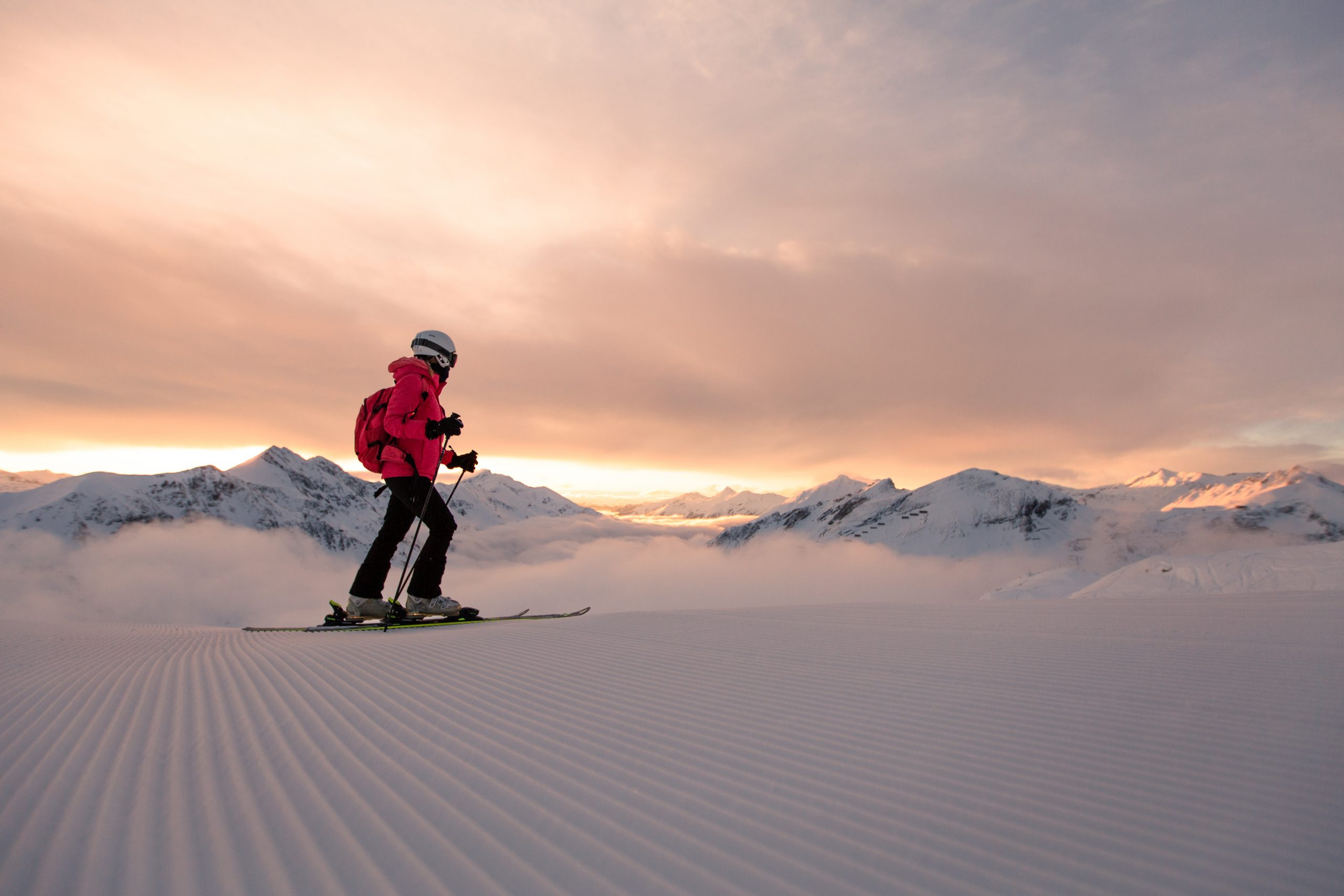 Person beim Skifahren bei Sonnenaufgang auf einer präparierten Schneepiste, mit Bergen und Wolken im Hintergrund.