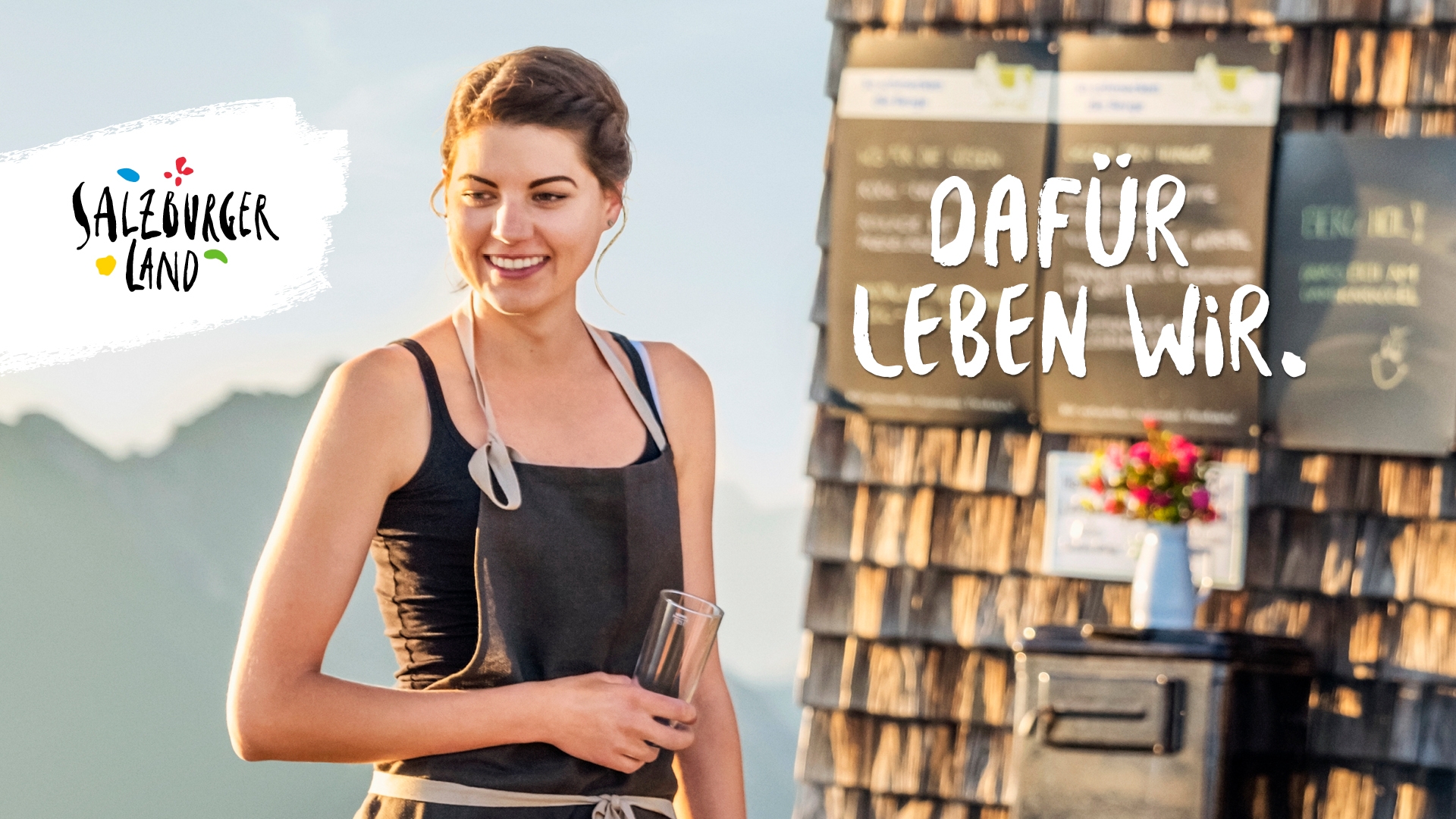 Lächelnde Frau in einer Schürze mit einem Glas in der Hand, daneben eine Berglandschaft und der Text Dafür leben wir.