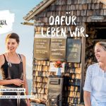 Zwei Frauen lächeln vor einer rustikalen Holzhütte in den Bergen, mit einem Salzburger Land Schild.