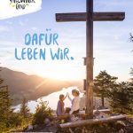 Zwei Personen sitzen an einem großen Holzkreuz auf einem Berg und blicken bei Sonnenuntergang auf einen malerischen See.