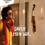 Ein Mann in einem gelben Pullover bewundert eine Geige, die aufrecht in einem Glaskasten steht.