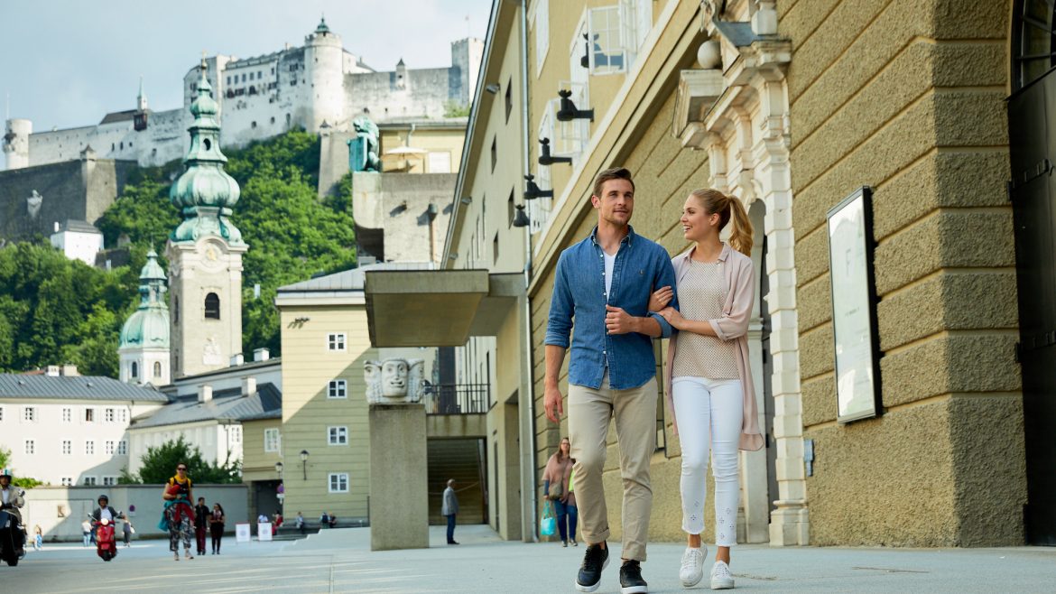Ein Paar spaziert gemeinsam auf einer Straße in der Stadt mit historischen Gebäuden und einer Festung im Hintergrund.
