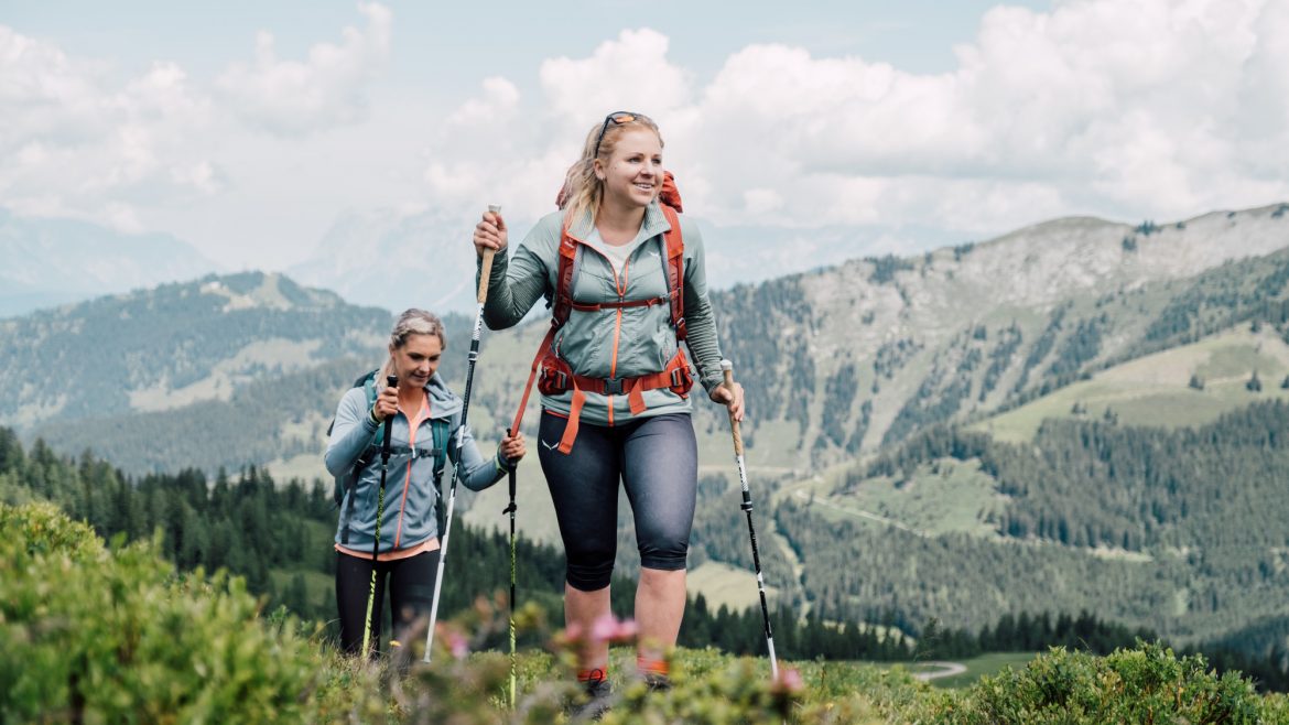 Zwei Frauen wandern mit Trekkingstöcken auf einem grünen Bergpfad mit malerischer Aussicht im Hintergrund.