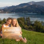 Zwei lächelnde Mädchen sitzen im Gras an einem See und halten ein Schild, das für das SalzburgerLand in Österreich wirbt.