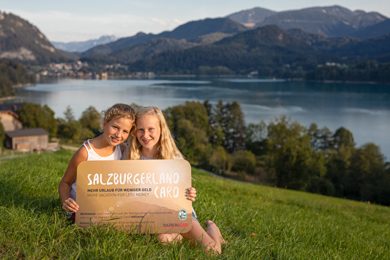 Zwei lächelnde Mädchen sitzen im Gras an einem See und halten ein Schild, das für das SalzburgerLand in Österreich wirbt.