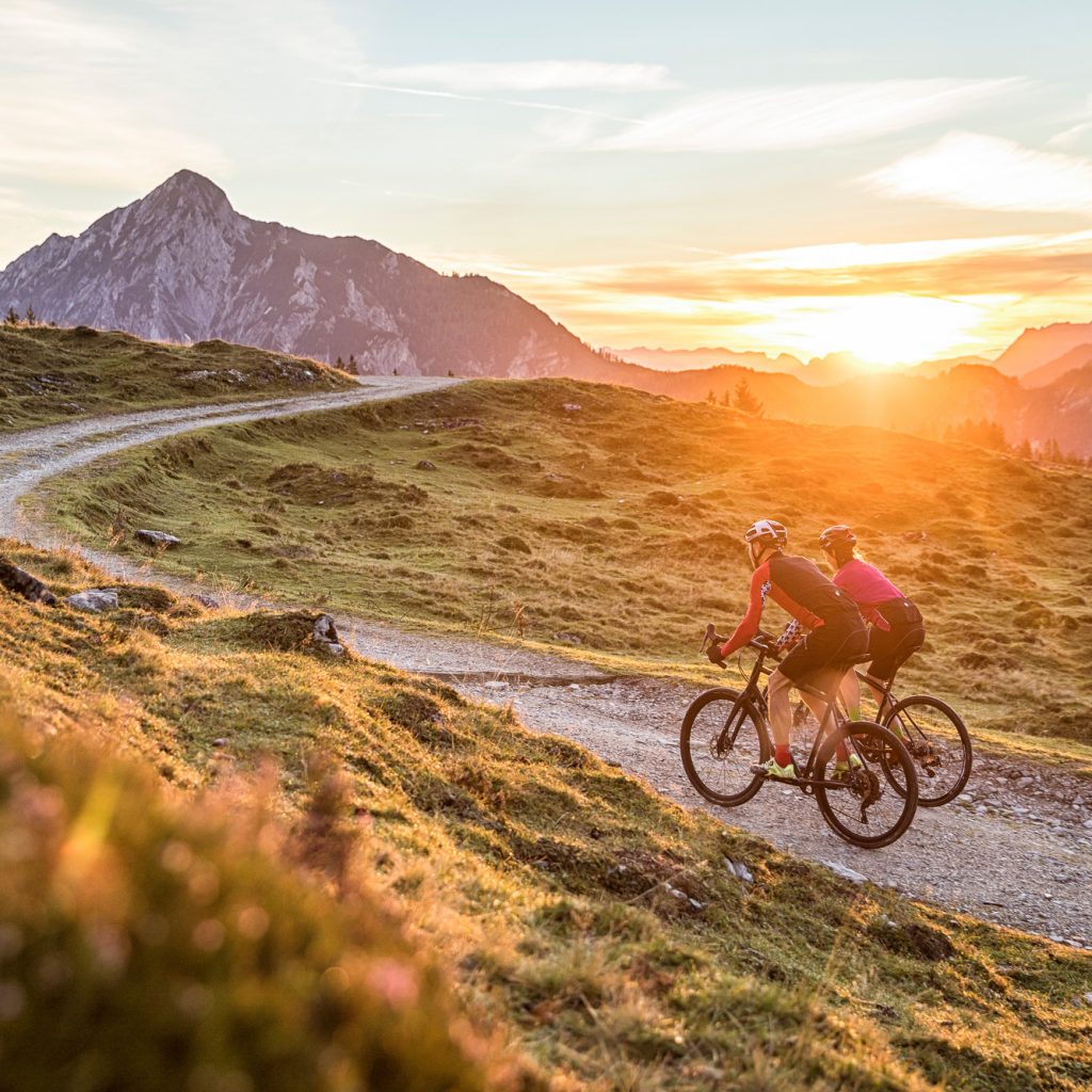 Zwei Radfahrer fahren bei Sonnenuntergang auf einem Bergweg mit malerischen Hügeln und Gipfeln im Hintergrund.