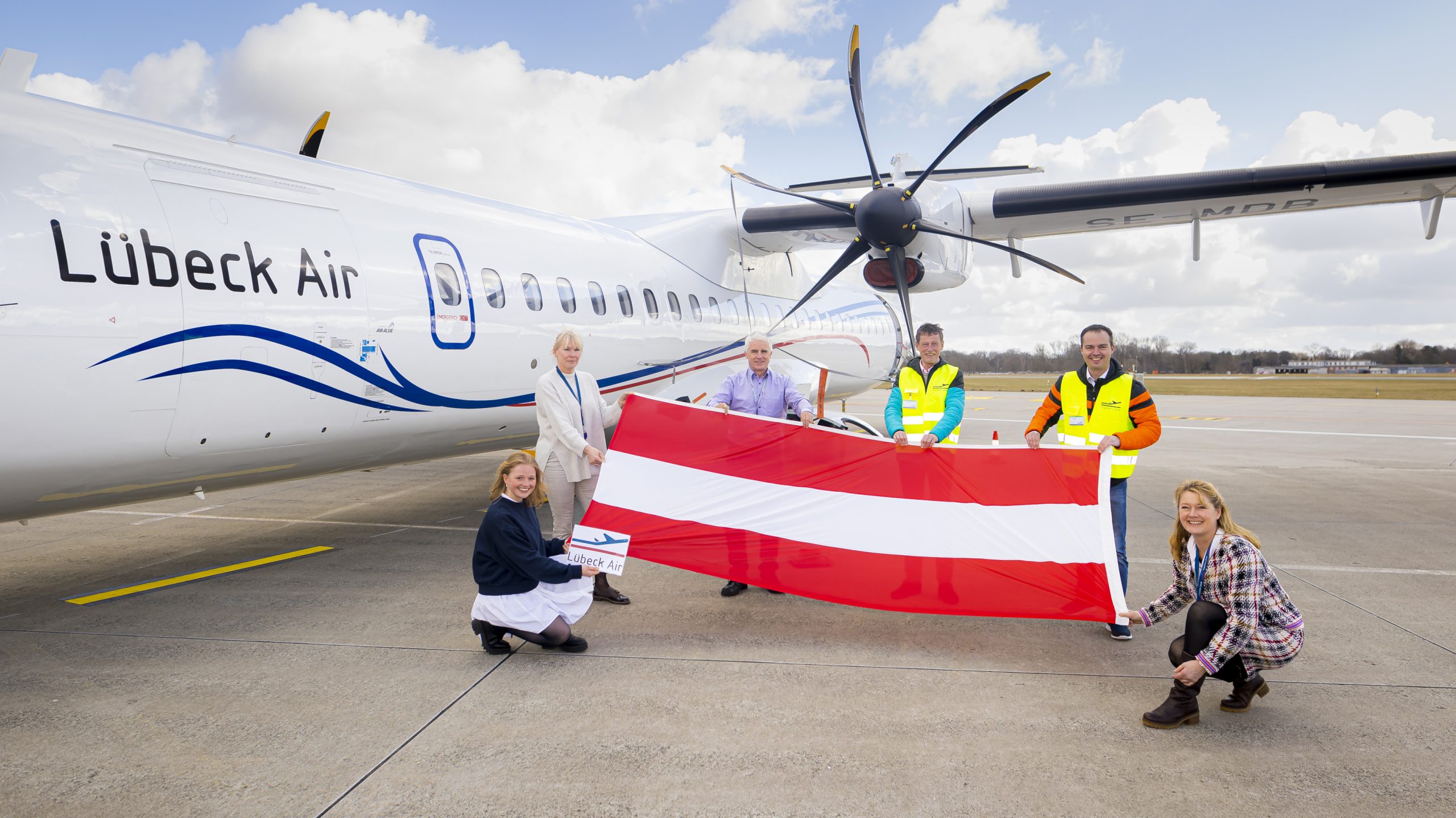 Sechs Personen halten eine österreichische Flagge vor einem Flugzeug der Lübeck Air auf dem Rollfeld des Flughafens.