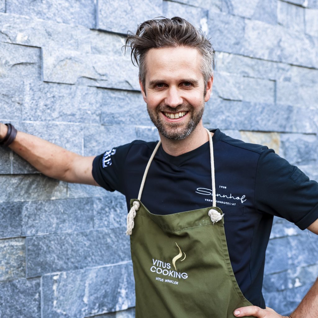 Ein lächelnder Mann in Schürze lehnt sich mit einem Arm an eine Steinmauer, im Freien.