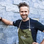 Ein lächelnder Mann in Schürze lehnt sich mit einem Arm an eine Steinmauer, im Freien.