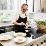 Ein Koch in einer gestreiften Schürze bereitet an einer modernen Küchentheke mit Tellern in der Nähe das Essen zu.