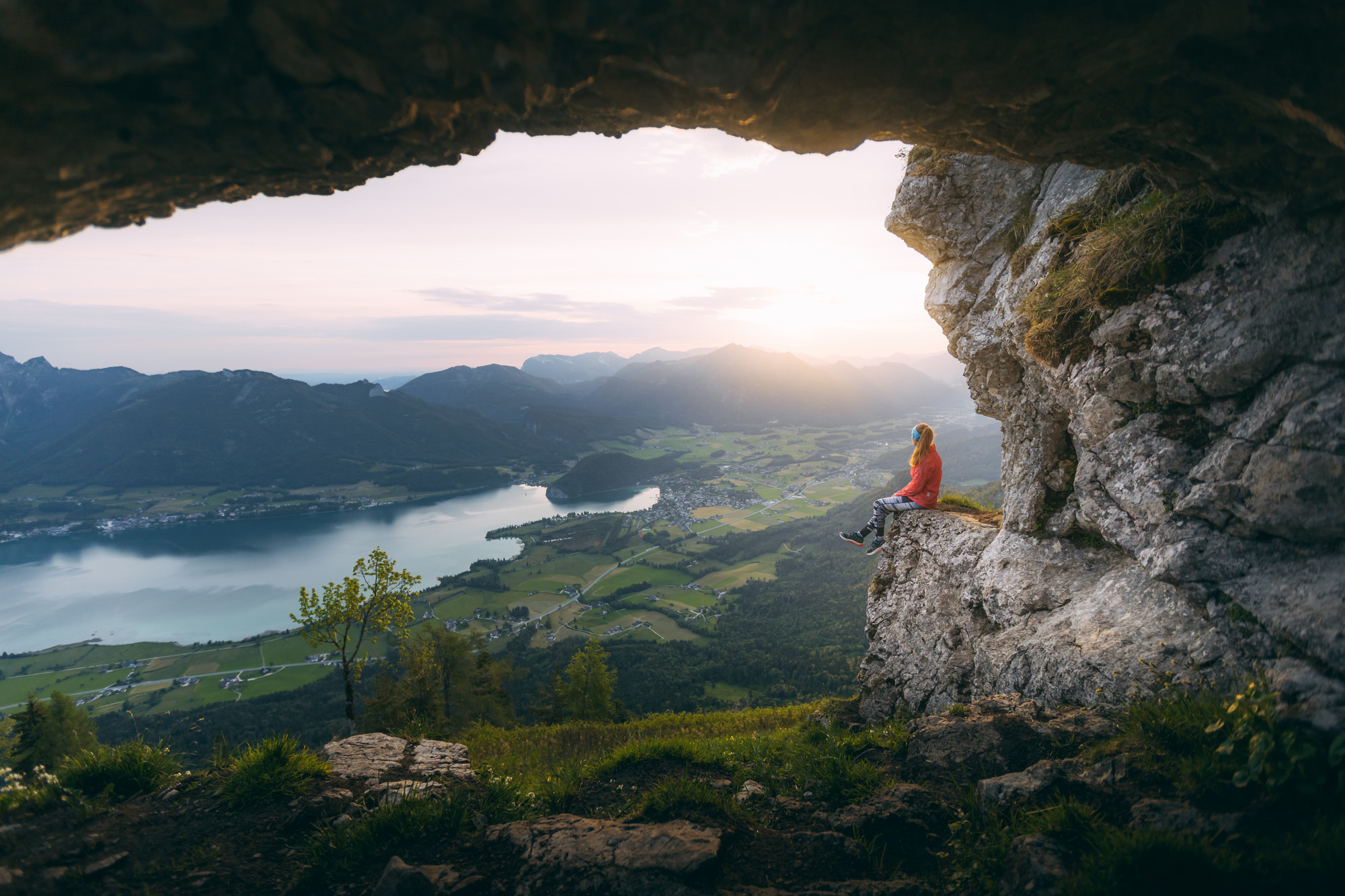 Eine Person sitzt auf einem Felsvorsprung mit Blick auf ein malerisches Tal, einen See und Berge bei Sonnenuntergang.