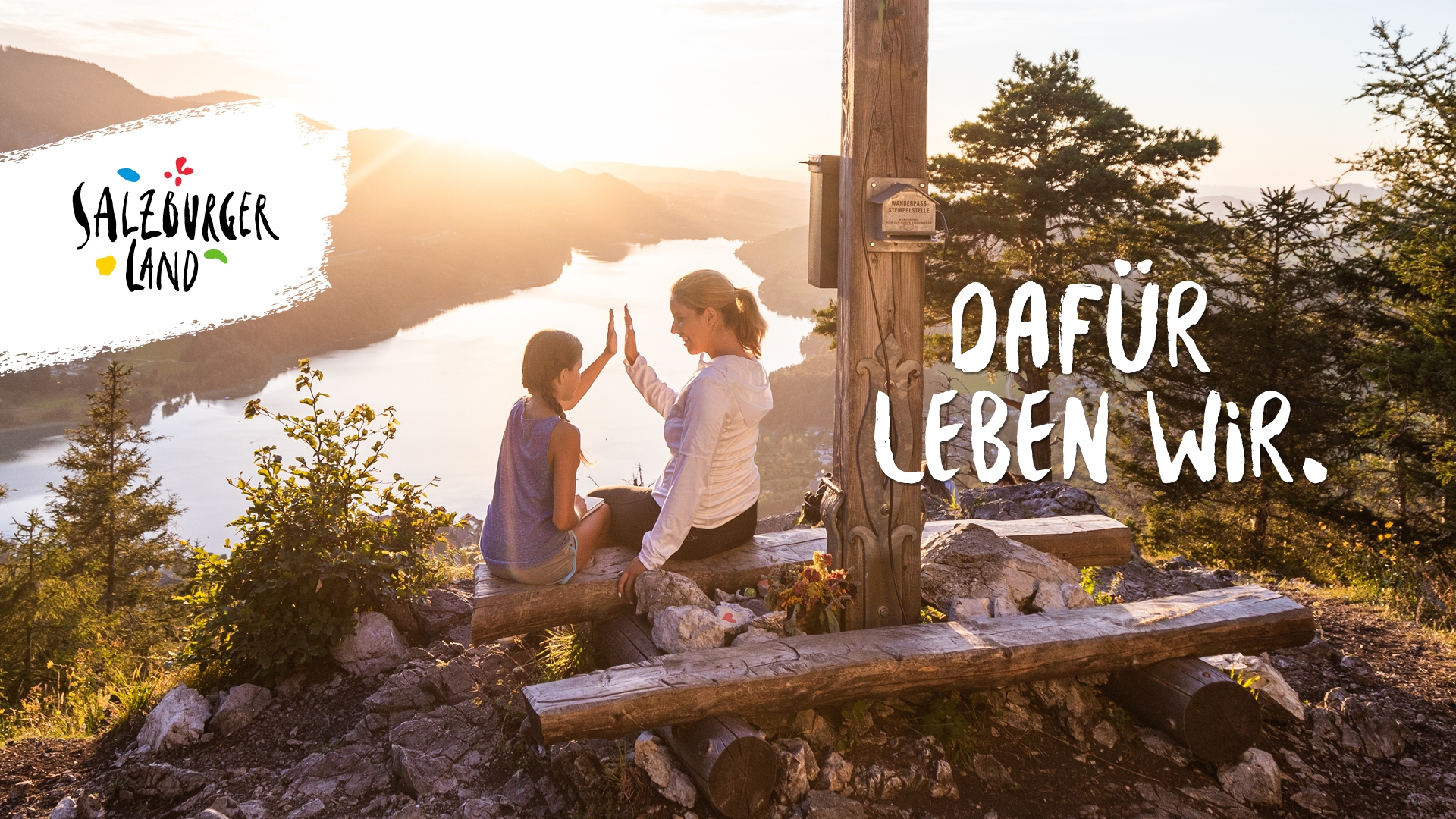 Eine Frau und ein Kind geben sich bei Sonnenuntergang an einem Holzkreuz auf einem Berg mit Blick auf einen See die Hand.
