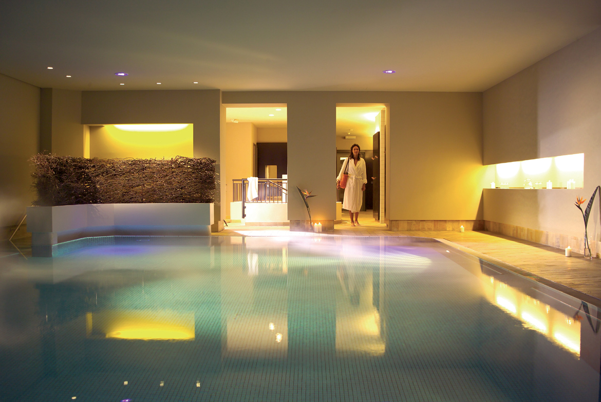 Eine Frau in einem weißen Bademantel betritt einen Indoor-Spa-Pool mit warmer Beleuchtung und modernem Dekor.