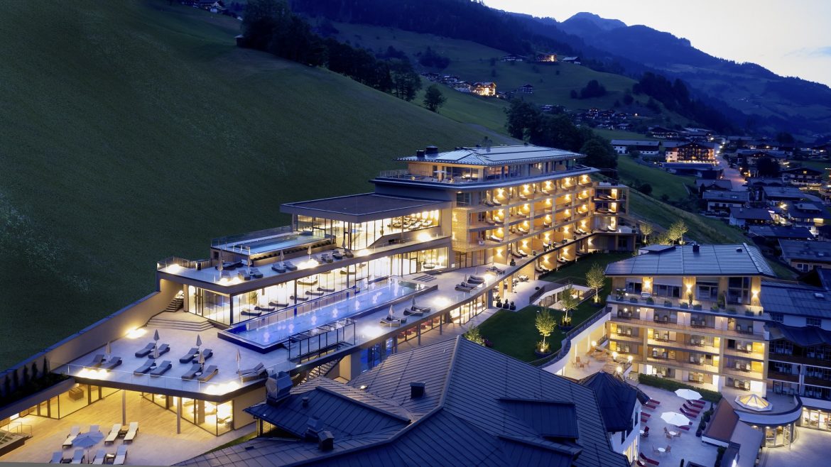 Modernes Hotel am Berghang in der Abenddämmerung mit leuchtenden Lichtern, Außenpool und Bergdorf im Hintergrund.