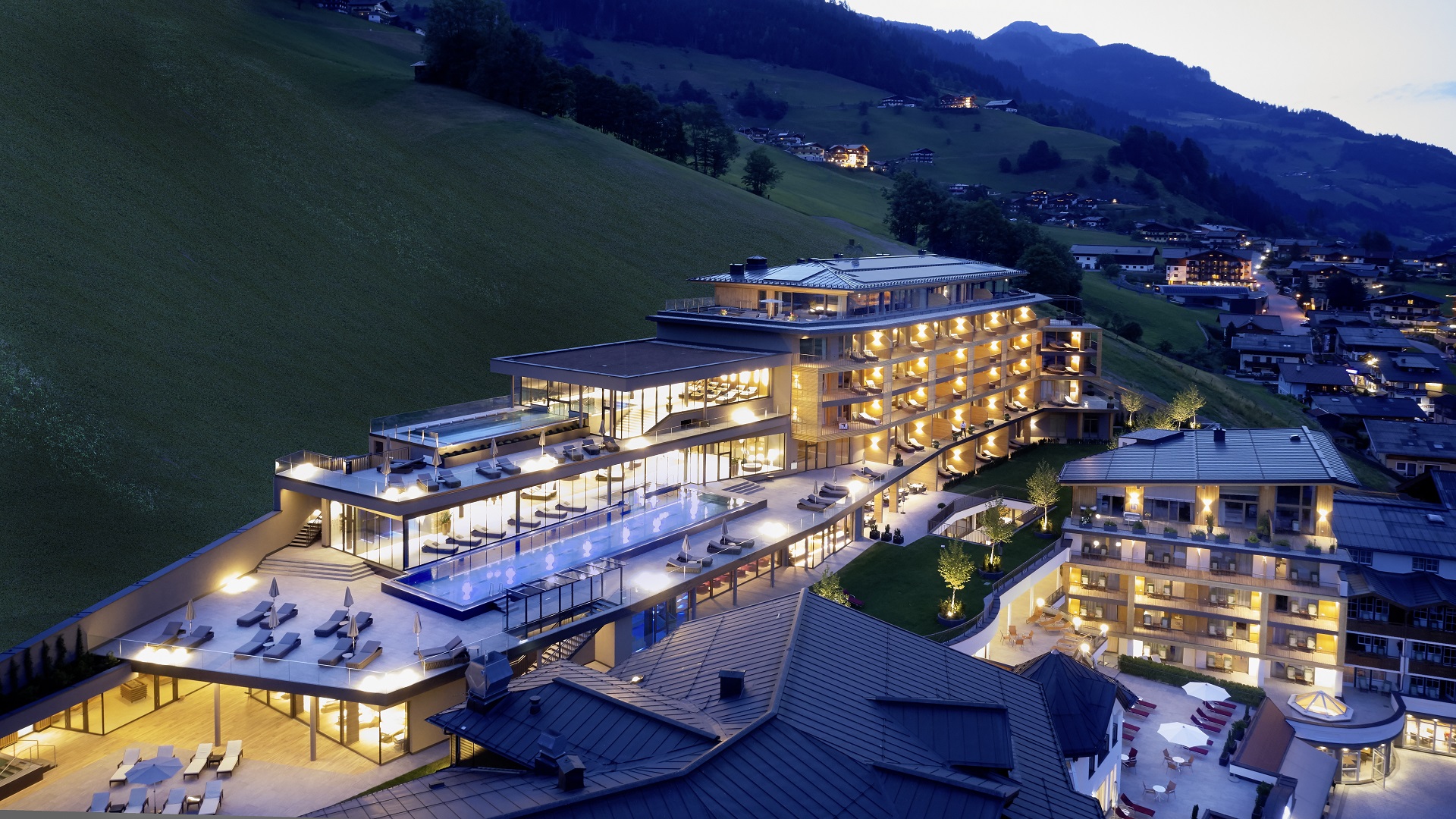 Modernes Hotel am Berghang in der Abenddämmerung mit leuchtenden Lichtern, Außenpool und Bergdorf im Hintergrund.