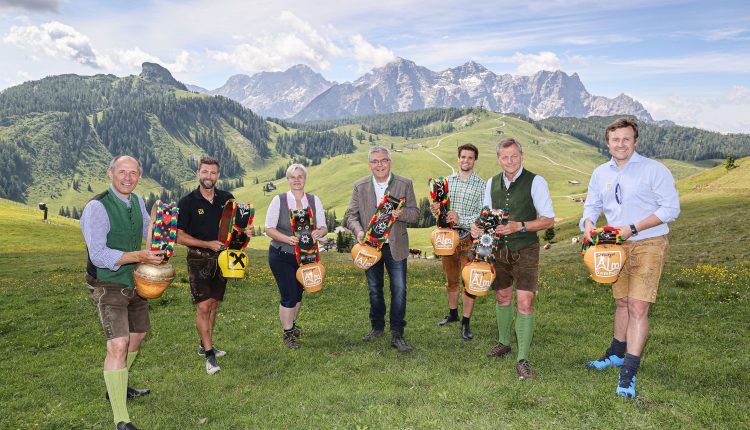 Acht Personen in traditioneller österreichischer Kleidung halten geschmückte Kuhglocken in einer grasbewachsenen Berglandschaft.
