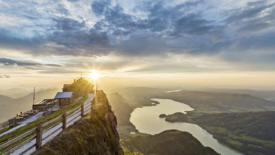 Die Sonne geht über einer Berghütte auf, mit Blick auf einen See und sanfte Hügel unter einem dramatischen, bewölkten Himmel.