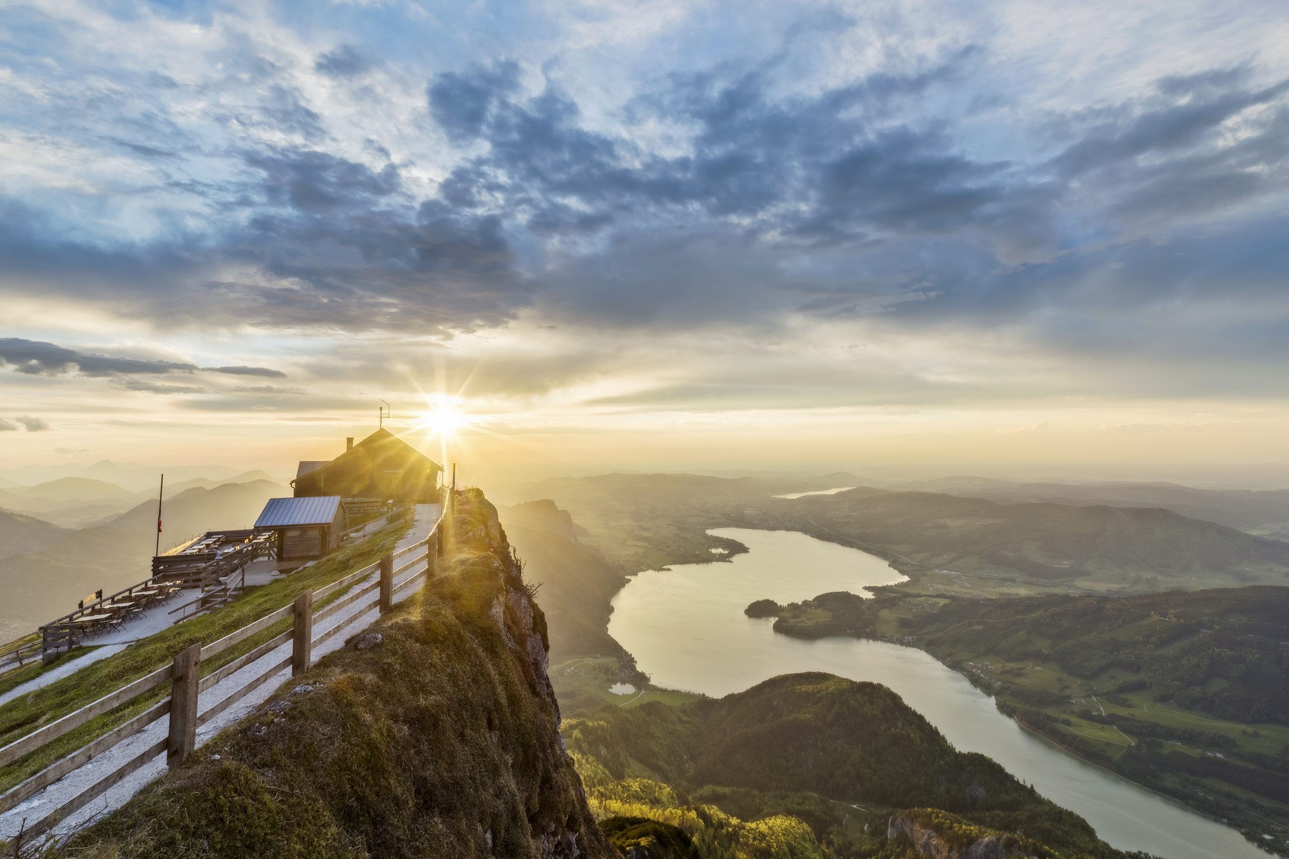 Die Sonne geht über einer Berghütte auf, mit Blick auf einen See und sanfte Hügel unter einem dramatischen, bewölkten Himmel.