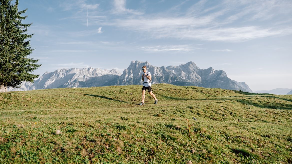 Person beim Wandern auf einem grasbewachsenen Hügel mit Bergen und blauem Himmel im Hintergrund.