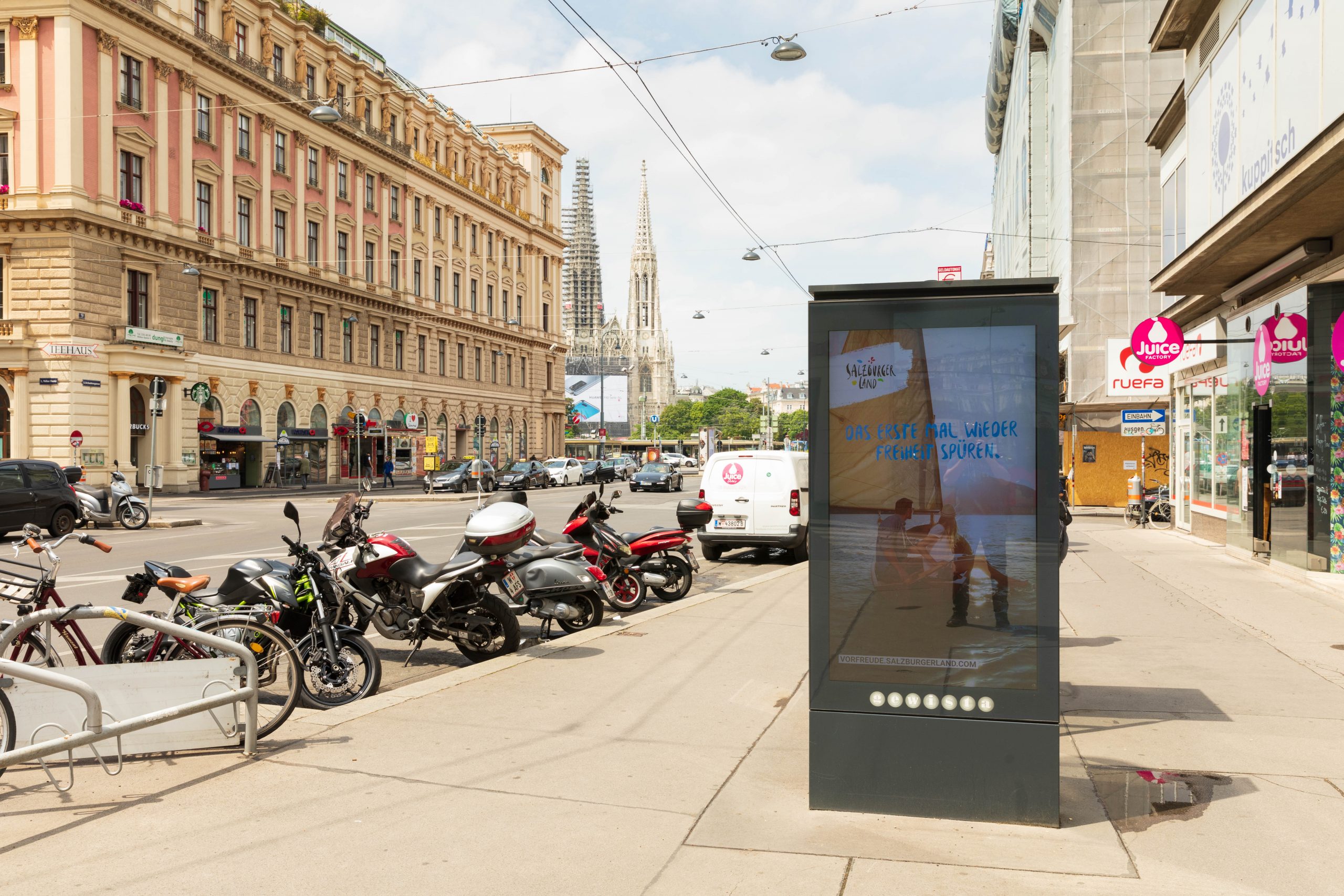 Eine Stadtstraße mit geparkten Motorrädern, Autos und einer digitalen Werbetafel; im Hintergrund historische Gebäude.