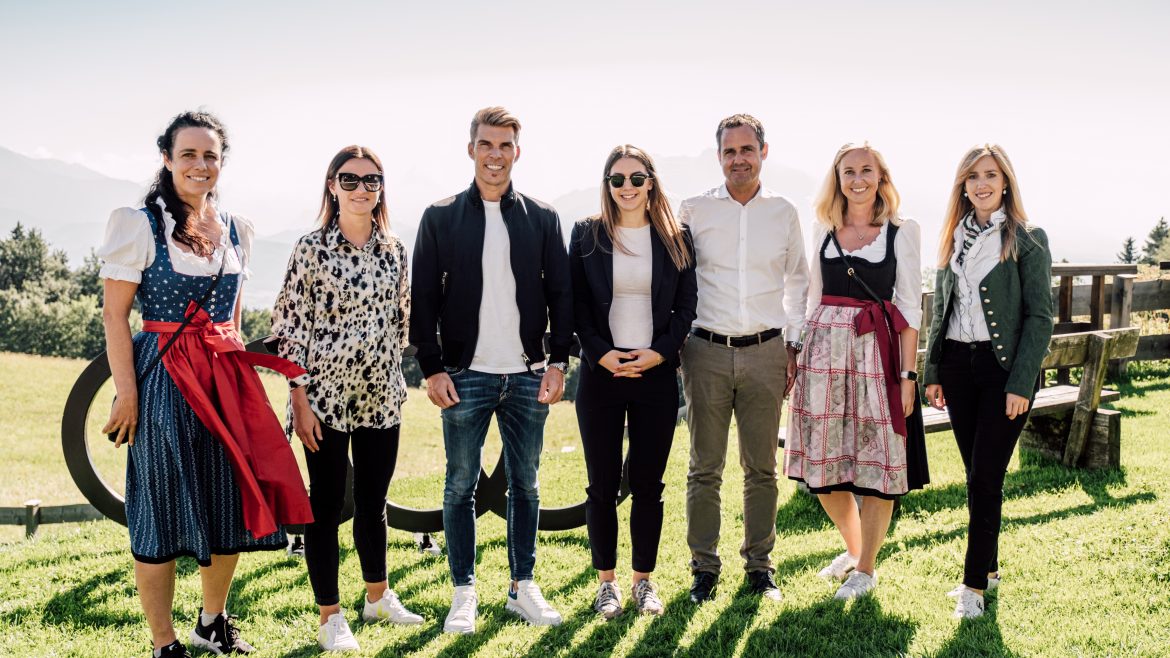 Sieben Menschen stehen lächelnd im Freien auf einer Wiese, mit Bergen im Hintergrund, einige in traditioneller österreichischer Kleidung.