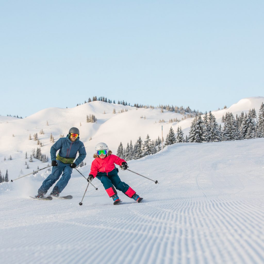 Erwachsener und Kind beim Skifahren auf einem sonnigen, verschneiten Berghang mit Bäumen im Hintergrund.