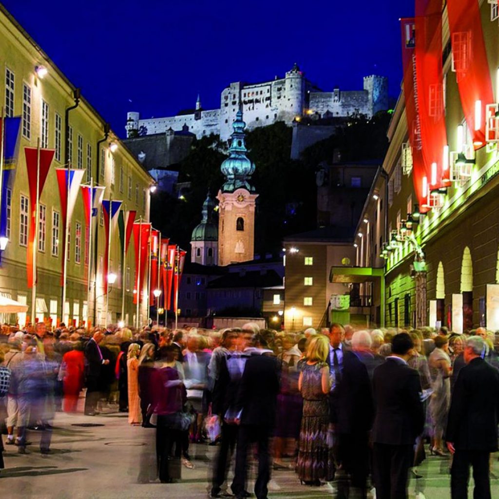 Feierlich gekleidete Menschen versammeln sich auf einer nächtlichen, festlich beleuchteten Straße, mit einem Schloss im Hintergrund.