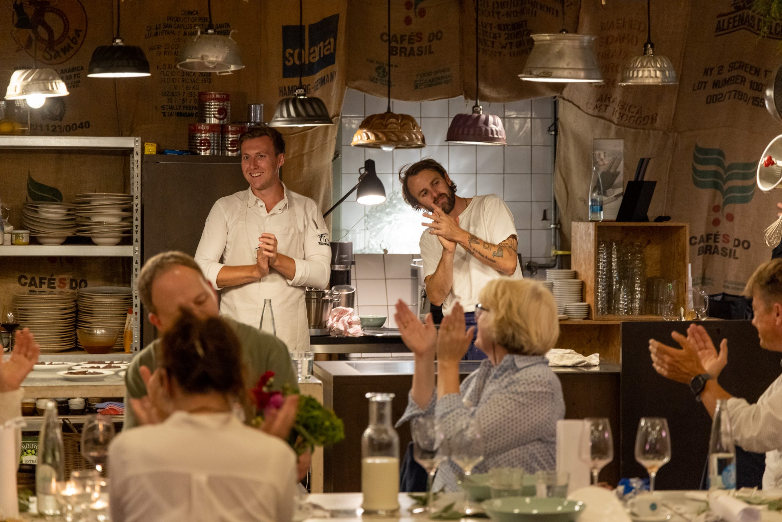 Zwei Köche stehen in einem warm beleuchteten, rustikalen Restaurant und lächeln, während die Gäste ihnen applaudieren.