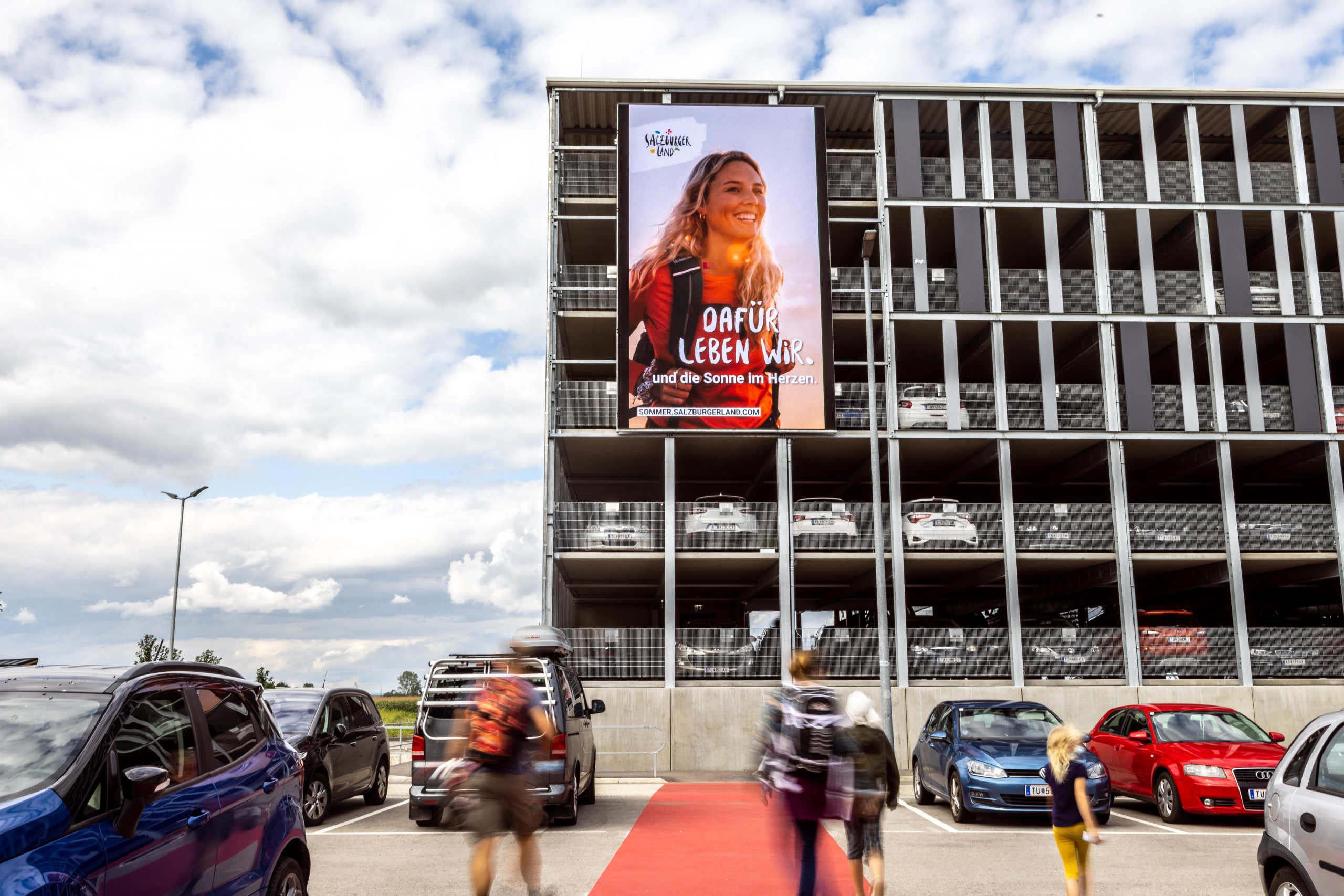 Menschen gehen in der Nähe eines Parkhauses vorbei, an dessen Fassade eine große Werbung mit einer lächelnden Frau angebracht ist.