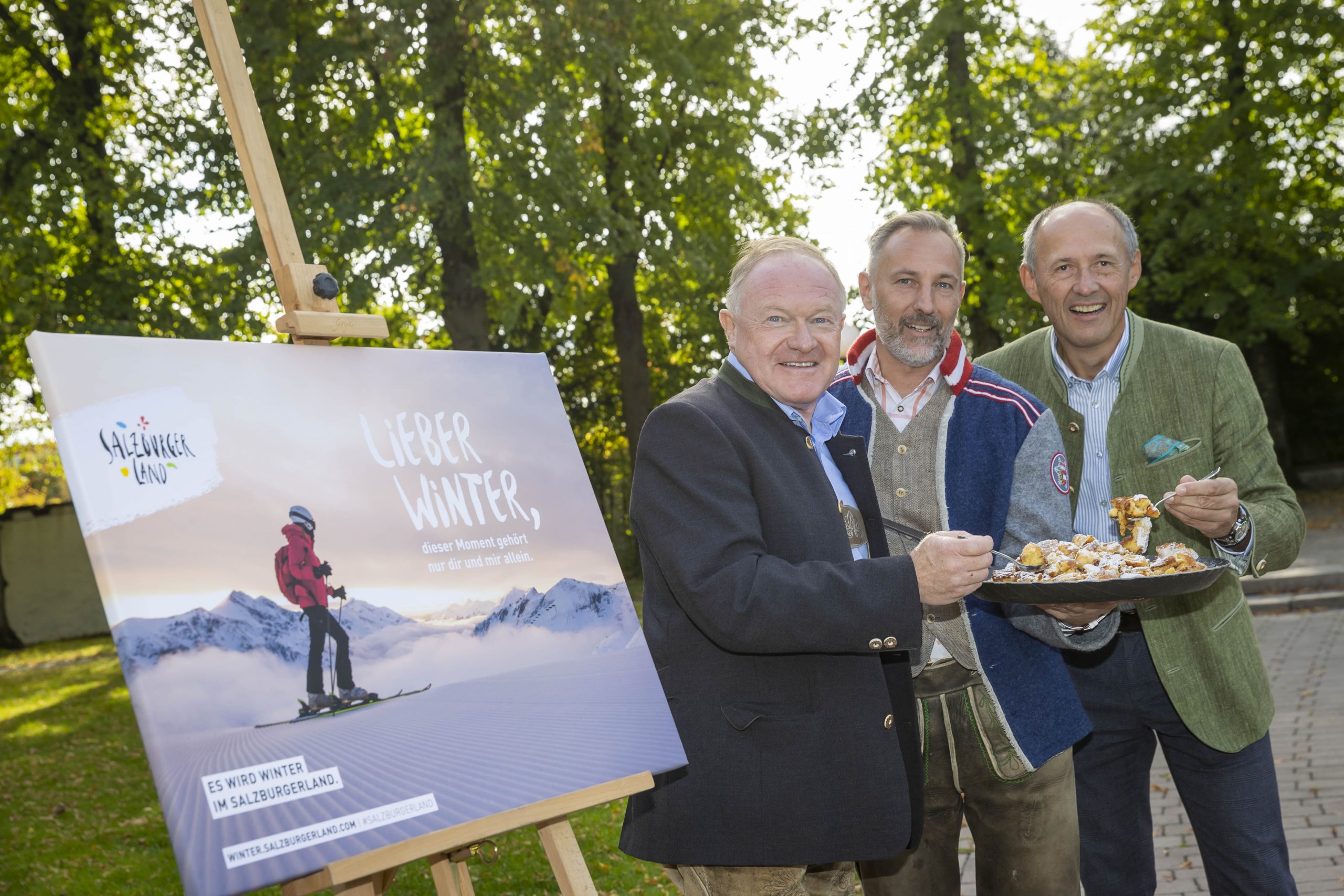 Drei Männer in Jacken stehen im Freien neben einem Poster eines Skifahrers, lächeln und halten einen Teller mit Essen in der Hand.