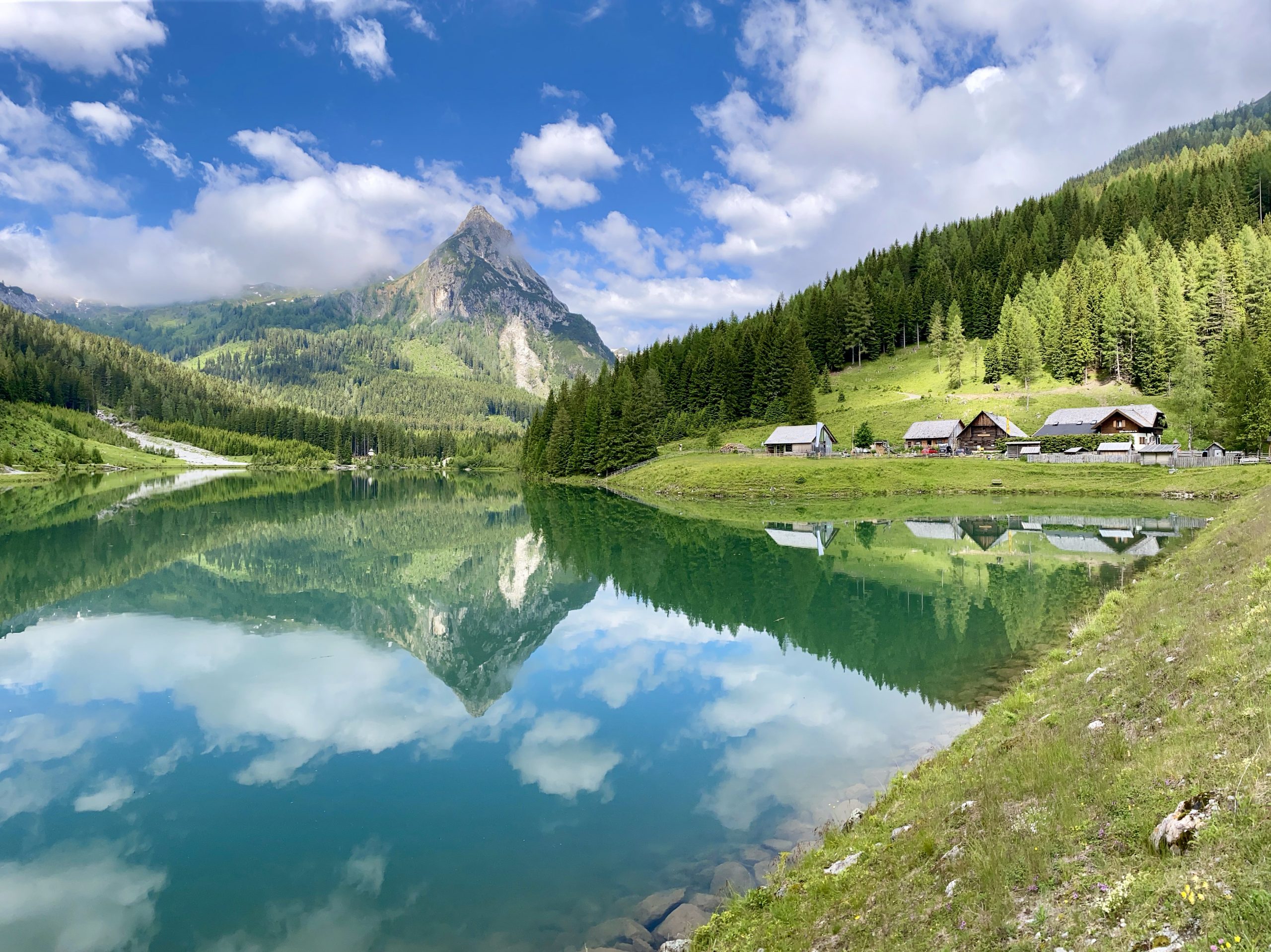 In einem ruhigen See spiegeln sich Berge, immergrüne Bäume und Hütten unter einem teilweise bewölkten blauen Himmel.