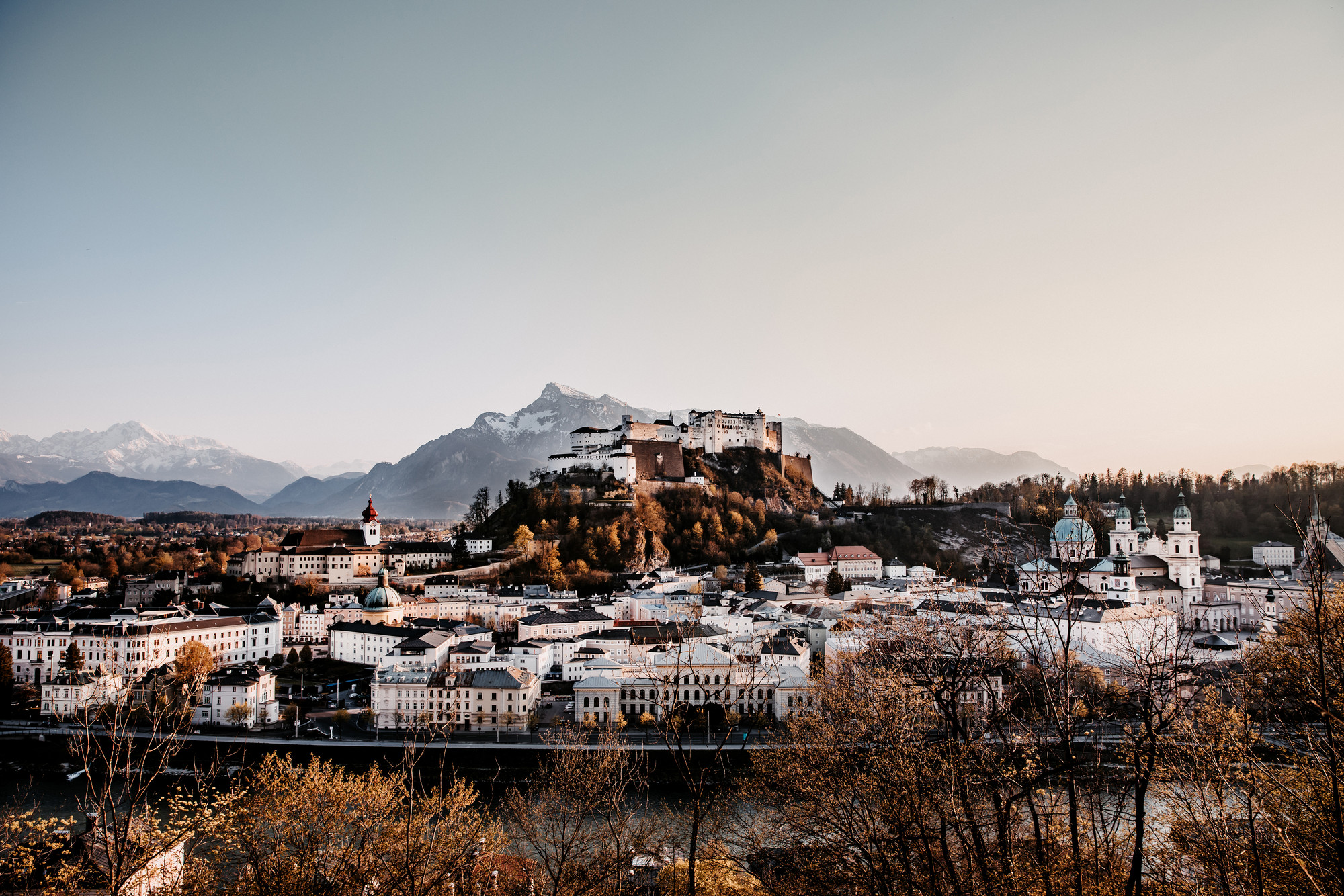 Ansicht von Salzburg, Österreich, mit der Festung Hohensalzburg auf einem Hügel und Bergen im Hintergrund bei Sonnenuntergang.