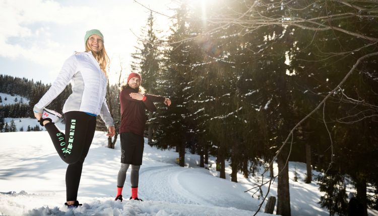 Zwei Personen strecken sich in Wintersportkleidung auf einem verschneiten Weg, umgeben von Kiefern und hellem Sonnenlicht.