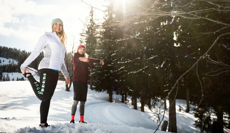 Zwei Personen strecken sich in Wintersportkleidung auf einem verschneiten Weg, umgeben von Kiefern und hellem Sonnenlicht.
