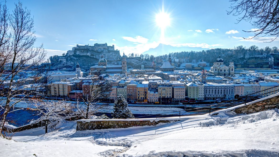 Schneebedecktes Stadtbild von Salzburg, Österreich, mit einer strahlenden Sonne und einer Festung auf einem Hügel im Hintergrund.
