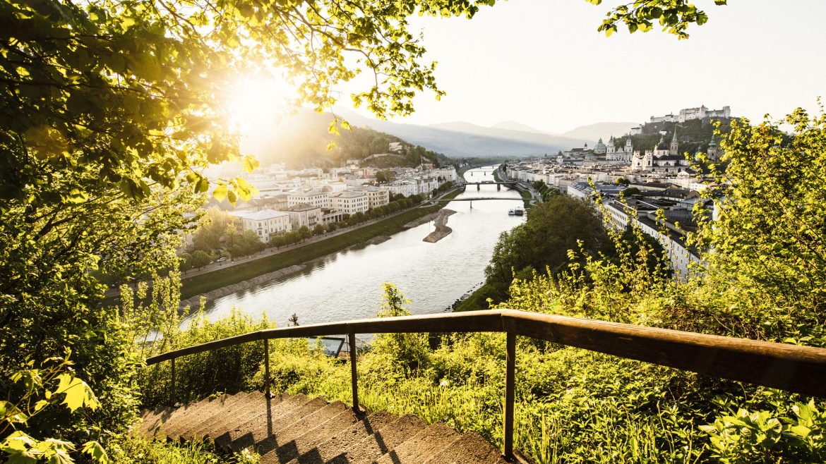Sonnenbeschienener Blick auf eine Stadt an einem Fluss, gesehen von einer begrünten Treppe mit Holzgeländer am Hang.